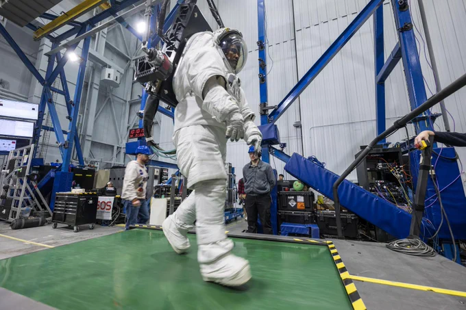 An image of Victor Glover wearing a white spacesuit walks on a treadmill-like platform inside a large testing facility while attached to a support system that helps simulate lunar gravity. Engineers and technicians observe nearby as the astronaut tests mobility and movement capabilities for future Artemis missions. Blue structural frames, monitoring equipment, and cables surround the test area.