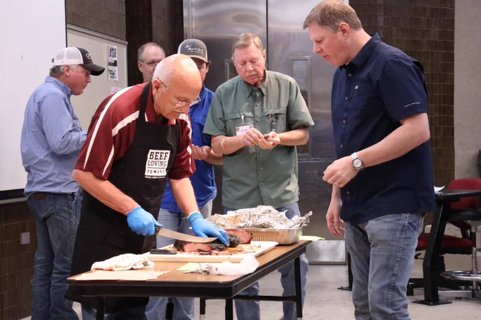 An individual wearing a "Beef Loving Texans" apron is slicing meat on a cutting board. Three others nearby are watching and tasting samples. A metal kitchen counter is in the background.
