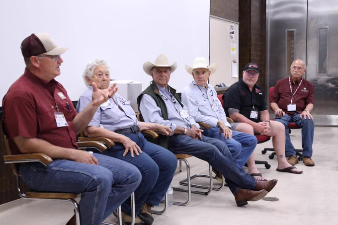 A group of six individuals, wearing casual and western-style attire, are seated in chairs during a panel discussion in a room. They are engaged in conversation, with one person speaking animatedly. The setting is informal, and they appear to be in a meeting or event space.
