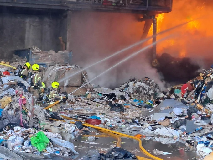 Three firefighters on the left of the image using hoses to spray water onto flames on the right of the image. The setting is a recycling centre.