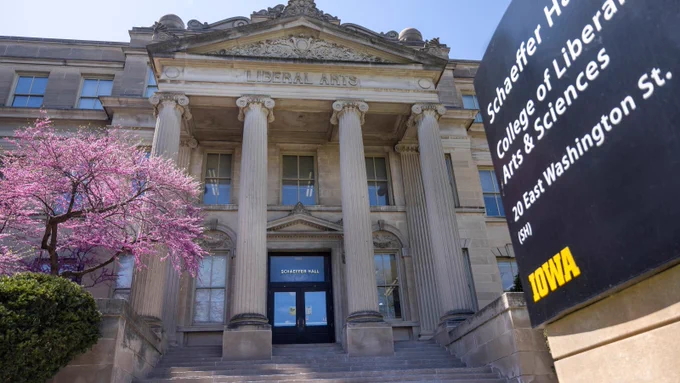 A photograph of Schaeffer Hall in the College of Liberal Arts and Sciences at the University of Iowa. A lilac tree blooms in front of the building, and a sign for the building, listing its address of 20 E. Washington St., is in the foreground.