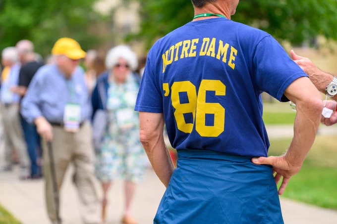 A person stands with hands on hips, wearing a blue shirt with "Notre Dame '86" printed on the back. In the background, several people are walking. The scene appears to be outdoors on a sunny day.
