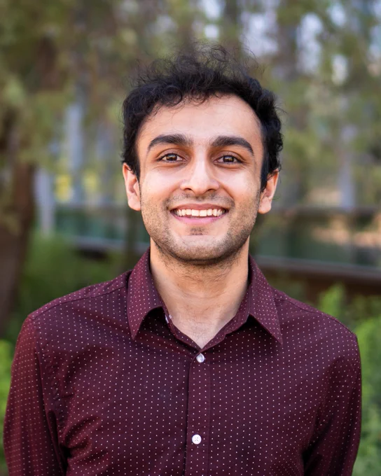 Belal Jamil smiles at the camera. He is wearing a maroon button-up shirt with a dotted-white pattern. Behind him are trees and foliage beside the windows of a nearby building. 