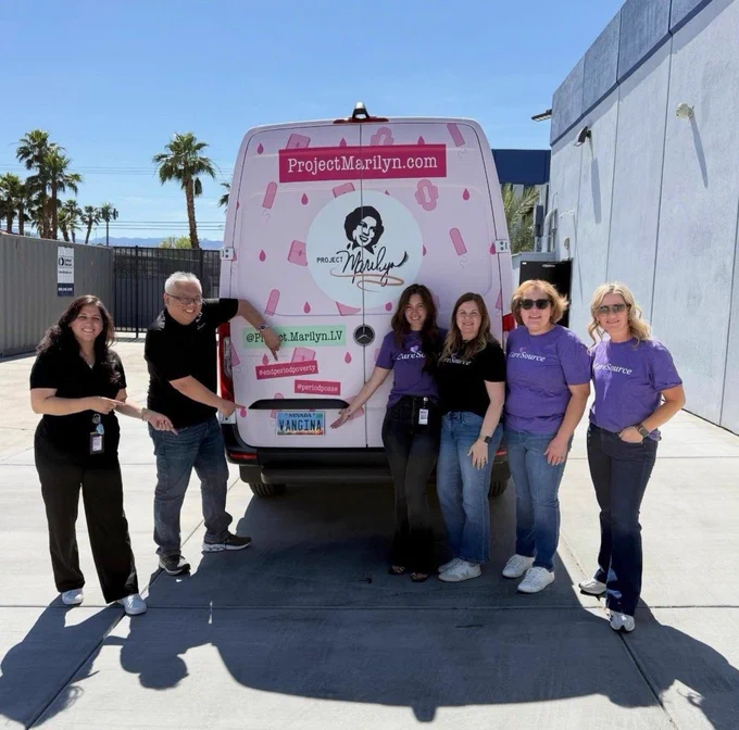 A group of six people is standing behind a van decorated with "Project Marilyn" branding. The van's rear door displays the website "ProjectMarilyn.com" and a graphic of the Project Marilyn logo. The individuals are smiling and wearing casual clothing. Palm trees and a building are visible in the background.