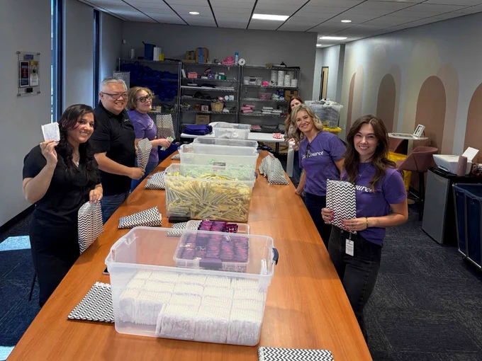 A group of people is standing around a long table in an office setting, packing items into patterned bags. The table is filled with clear bins containing various items. Some individuals are wearing purple shirts with a recognizable logo. Shelving with more supplies is visible in the background.
