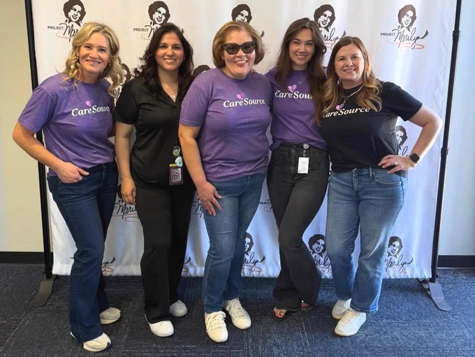 A group of five people stands together, smiling in front of a "Project Marilyn" backdrop. Three individuals wear purple "CareSource" T-shirts, one wears a black top, and another wears a black "CareSource" T-shirt. All are dressed casually with jeans.