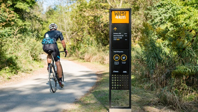 Cyclist riding on a paved trail surrounded by dense green foliage with a black and yellow trail sign.