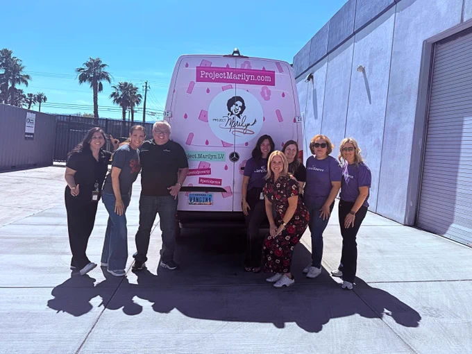 A group of people smiling and standing in front of a van with "Project Marilyn" branding in a sunny outdoor setting. The van is parked near a large building and palm trees are visible in the background.