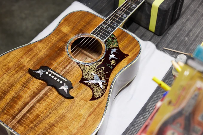 A detailed close-up of a beautifully crafted Gibson Doves in Flight acoustic guitar lying on a table. The guitar features intricate inlay work near the soundhole, depicting birds and foliage. The wood displays a rich, warm tone with a polished finish.