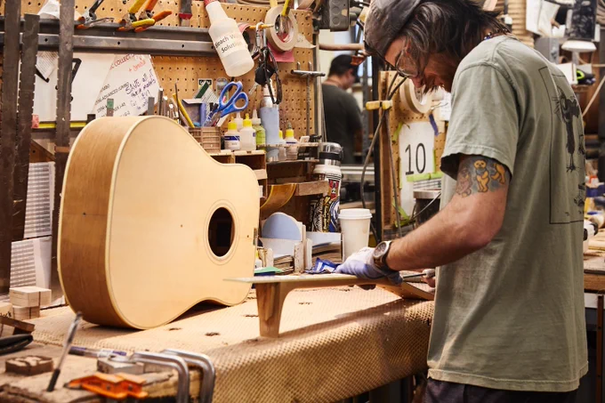 A person is working on a Gibson acoustic guitar in a workshop filled with tools and materials. The workspace appears organized, with various instruments and supplies around, including a pegboard with items hanging and a workbench with the guitar resting on it.