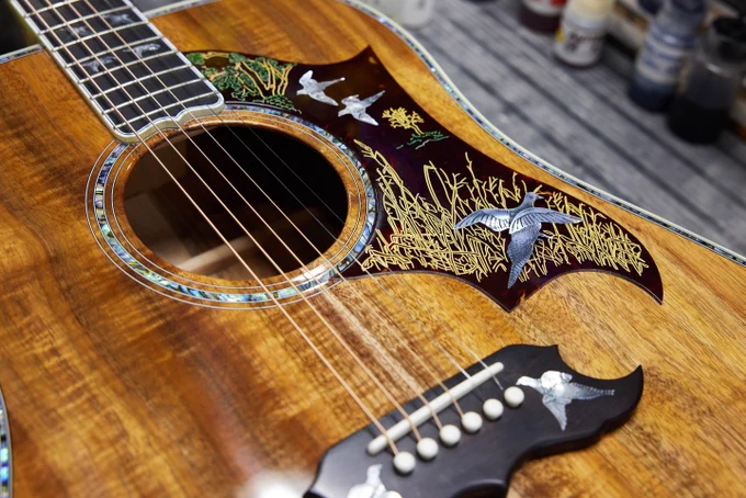 This image shows a close-up of a wooden Gibson Doves in Flight acoustic guitar with intricate inlays around the soundhole and on the pickguard. The inlays depict birds and foliage in silver and gold colors. The guitar's polished wood surface highlights its craftsmanship.