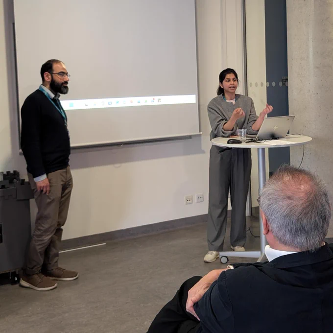Two people presenting at the front of a room; one speaks beside a laptop on a small round table while another stands nearby, with a projection screen behind and an audience member in the foreground.