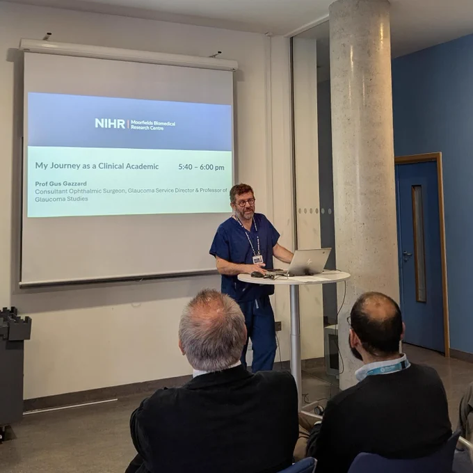 A speaker in medical scrubs presents beside a laptop and projected slide reading “NIHR Moorfields Biomedical Research Centre – My Journey as a Clinical Academic,” with seated audience members in the foreground.