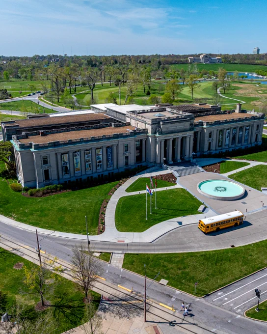 An aerial of the Missouri History Museum’s north entrance shows pathways and roadways crisscrossing in the foreground as a landscape of trees and foliage expand underneath a blue sky behind the rectangular building. 