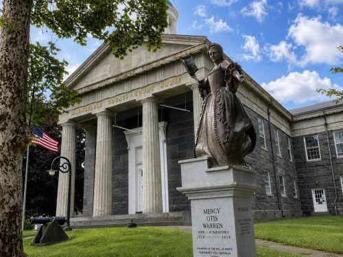 Statue of Mercy Otis Warren stands before Barnstable County Courthouse, with Greek Revival architecture, American flag, trees, and blue sky.