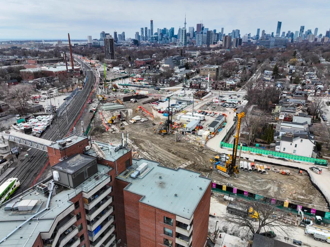 Aerial view of a large construction site near a cityscape in Toronto. The site is surrounded by residential buildings and train tracks, with various construction machinery visible. The Toronto skyline is in the background, featuring the CN Tower and other high-rise buildings. 