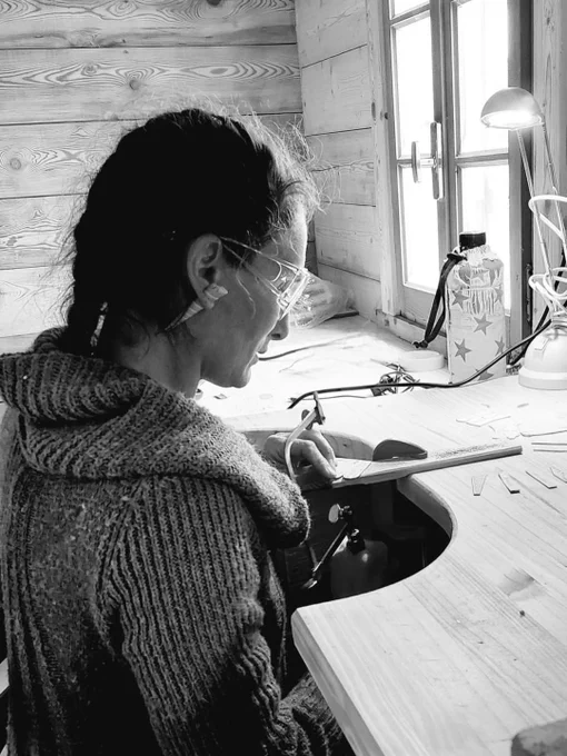A black and white image of a person wearing glasses, concentrating on crafting at a wooden workbench in a cozy workspace. The scene is serene and focused, highlighting the artisan's dedication.