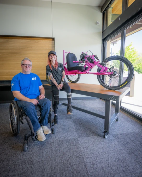 Two people, one in a wheelchair, pose indoors beside a bright pink adaptive handcycle displayed on a table by tall windows.