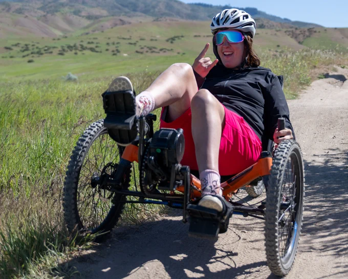 Person wearing a helmet and mirrored sunglasses rides an orange recumbent trike on a dirt trail through grassy hills.