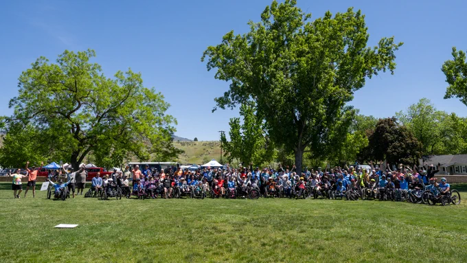 Large group of people, many using adaptive bikes and wheelchairs, gathered on a sunny park lawn beneath tall trees.
