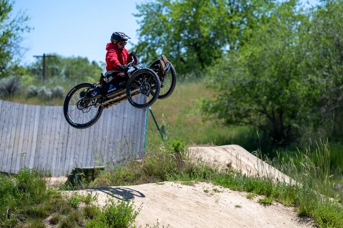 Rider in a red hoodie and helmet launches a three-wheeled handcycle off a dirt jump on a grassy trail.