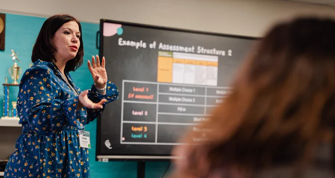 A woman, animatedly speaking, stands front of a group next to a TV showing slides.