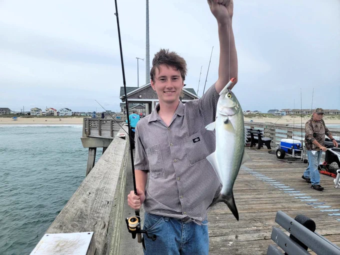 A person on a pier holds up a fish they caught, smiling. They are holding a fishing rod in the other hand. The background shows the ocean and a line of beach houses.