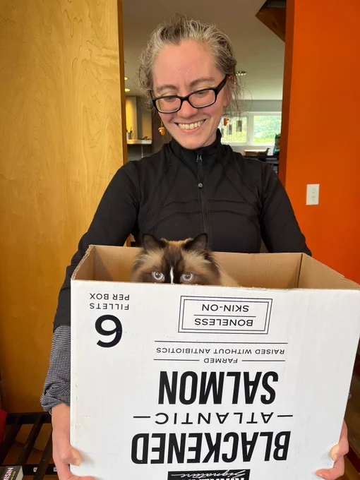 A photo of me smiling down while holding a box. My cat Biscuit pokes his head out of the box, glowering at his domain, and expressing disappointment that the box which says "blacked salmon" contained no salmon (just him)