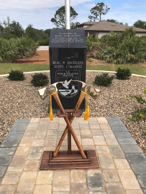Memorial monument dedicated to Beau W. Sauselein and Scott J. Maness of the U.S. Fish & Wildlife Service, featuring a plaque and crossed pulaskis on a paved platform.