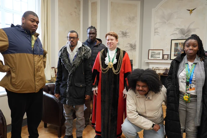 A group of six people, including a man in mayoral robes, pose for a photo in a room.