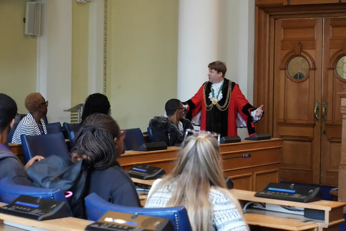A man in ceremonial robes speaks to an audience in a council chamber.