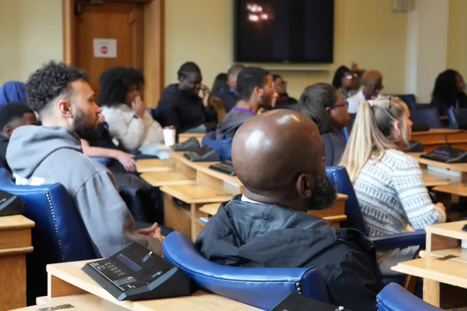 A diverse audience seated in a lecture hall, with a focus on the back of a bald Black man in the foreground.