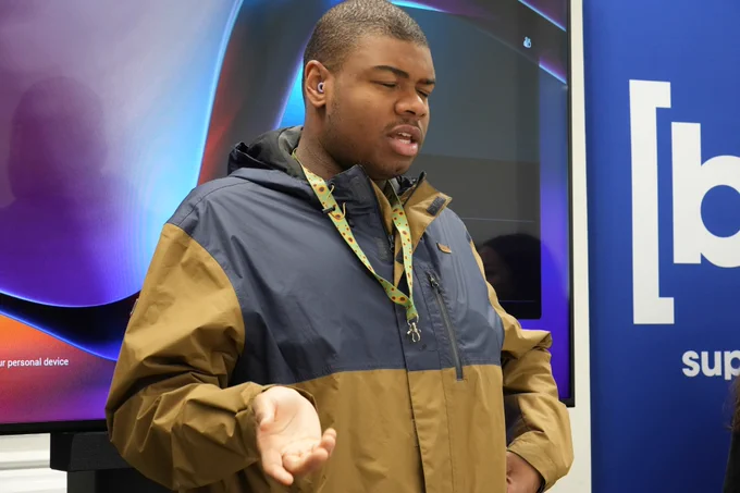 A young Black man in a brown and blue jacket gestures with his hand, standing before a large screen.