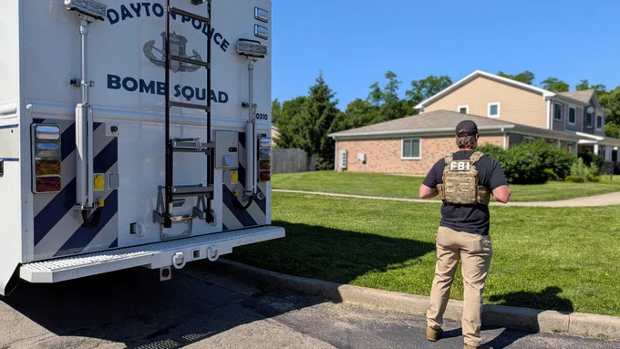 FBI agent wearing tactical gear stands near a Dayton Police Bomb Squad vehicle in a suburban neighborhood.