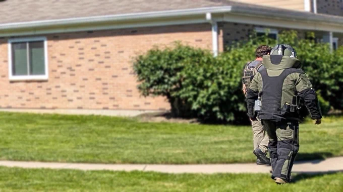 A person in a bomb disposal suit walking on grass toward a brick house with another individual.