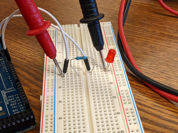Close-up photo of red and black multimeter probes touching jumper wires plugged into a solderless breadboard, showing a hands-on continuity or voltage check during circuit debugging.