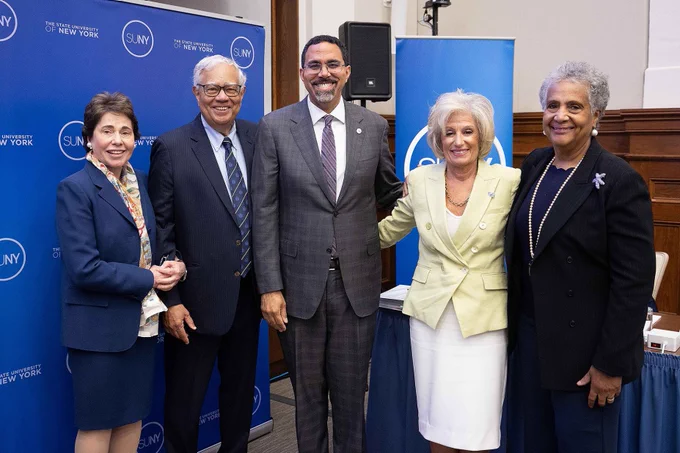 Five smiling adults, two men and three women, pose in front of blue \"THE STATE UNIVERSITY OF NEW YORK SUNY\" banners. From left: woman in navy suit, man in dark suit, man in grey suit, woman in yellow jacket & white skirt, woman in black jacket & pearl necklace.