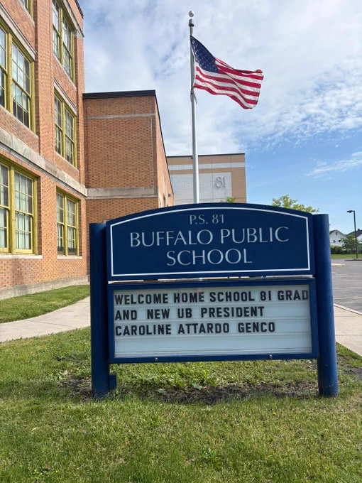 A blue sign for P.S. 81 Buffalo Public School stands on a grassy lawn. Its message board reads: 'WELCOME HOME SCHOOL 81 GRAD AND NEW UB PRESIDENT CAROLINE ATTARDO GENCO'. A brick school building with \"81\" visible, an American flag, and a cloudy sky are in the background.