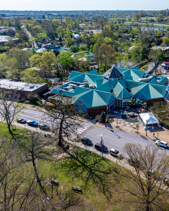 An aerial over the Saint Louis Zoo's north entrance has an expanded horizon of full-leaf trees behind the multi-sloped angels of it's visitor center building.
