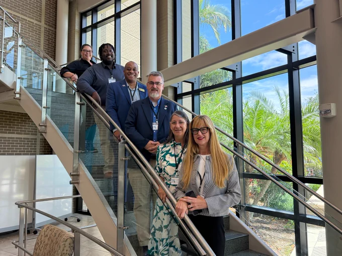 A group of six people standing on a modern staircase in a well-lit building with large windows. The view outside shows palm trees and a blue sky. They are dressed in formal attire and smiling at the camera.