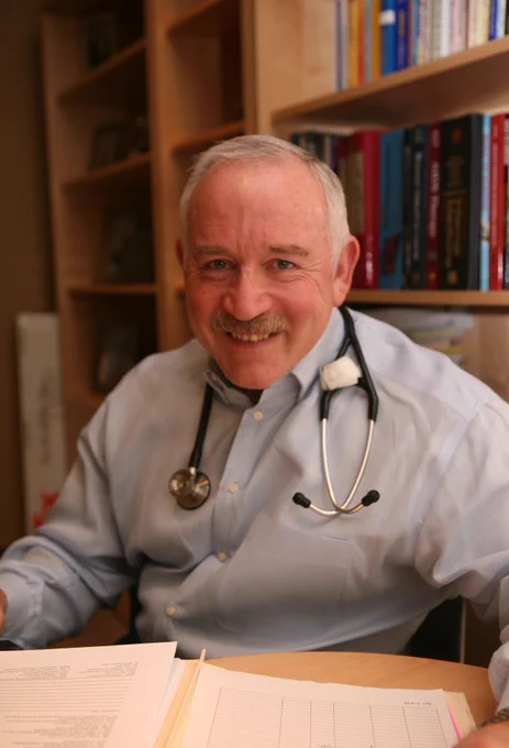 Dr. Brady Allen sitting at a table with papers in front of him and bookcases behind him. He is wearing a blue shirt and stethoscope.