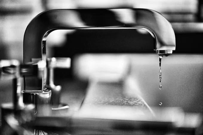 Close-up of a modern faucet with a single water droplet falling, captured in black and white.