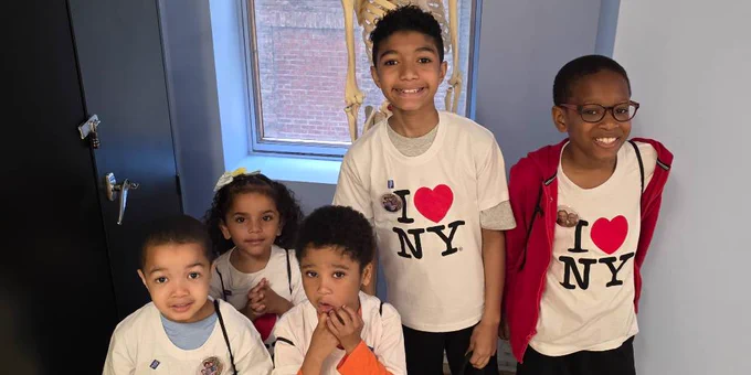 Five children wearing "I ♥ NY" shirts stand indoors in front of a window with a skeleton model behind them.