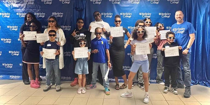 Group of children and adults standing in front of a Berkeley College backdrop, holding certificates, celebrating an achievement event.