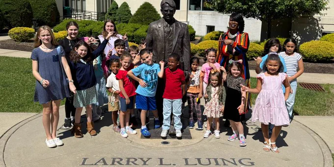 Group of children and an adult posing around a Larry L. Luing statue outside a building on a sunny day.
