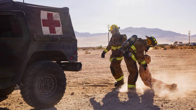 Aircraft rescue firefighters drag a simulated casualty away from a military vehicle during emergency response training in a desert environment.