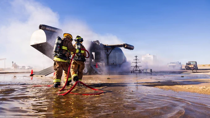 Two firefighters advance a hose line through standing water after extinguishing a simulated aircraft fire.