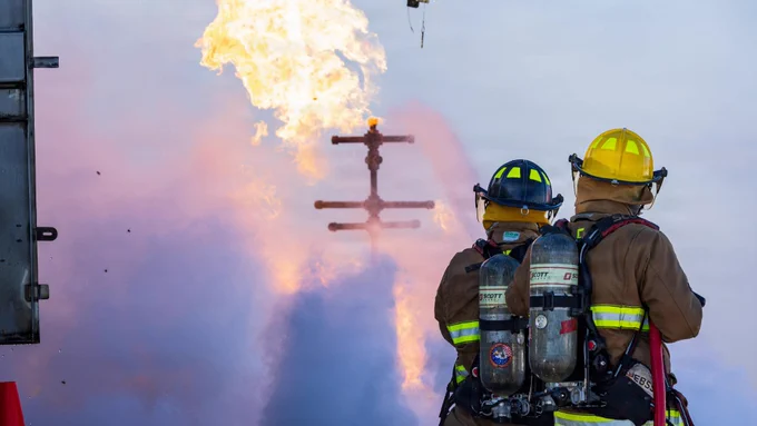 Two firefighters direct water onto a live-fire training prop while flames and steam rise around them.