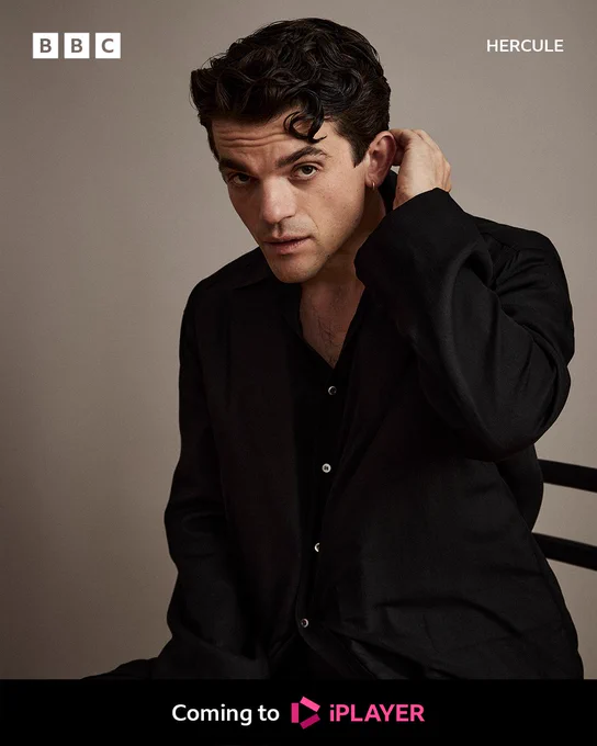 A head and shoulders shot of a man with dark hair in a dark buttoned shirt, looking to camera with one hand reaching around to the back of his neck as he sits on a chair