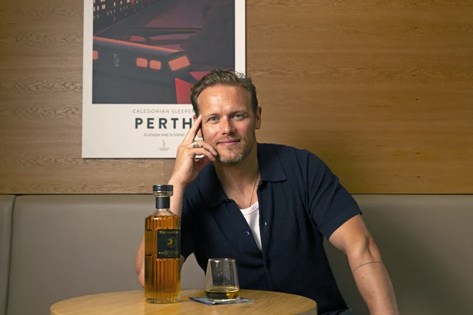Sam Heughan sitting at a round wooden table with a glass of whiskey and a bottle, resting his head on his hand.